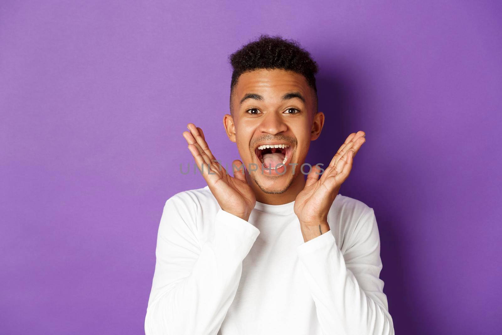 Close-up of excited african-american man, scream of joy and looking amazed at camera, hear good news, standing over purple background.