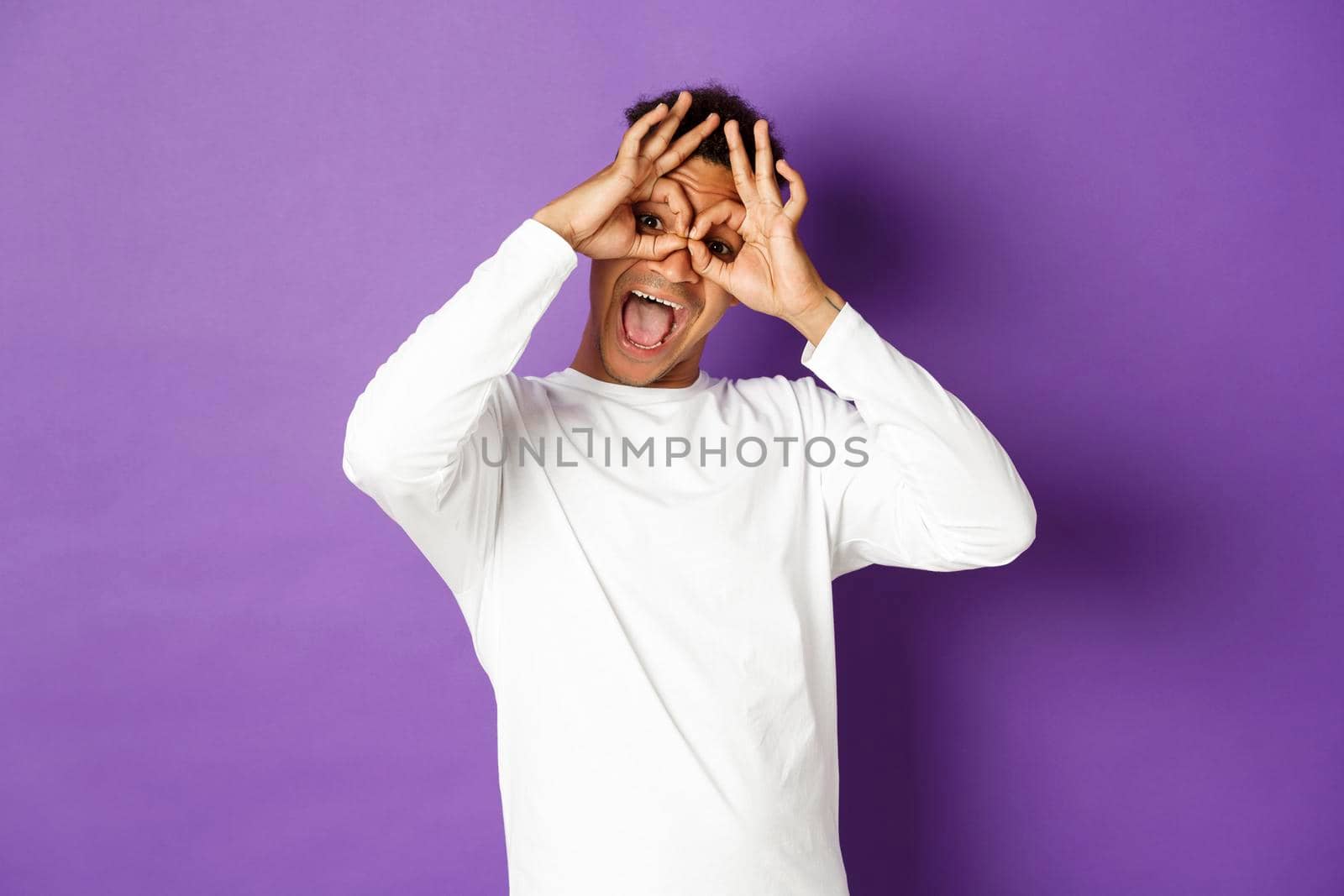 Image of funny and happy african-american man in white sweatshirt, grimacing and making faces, standing over purple background.