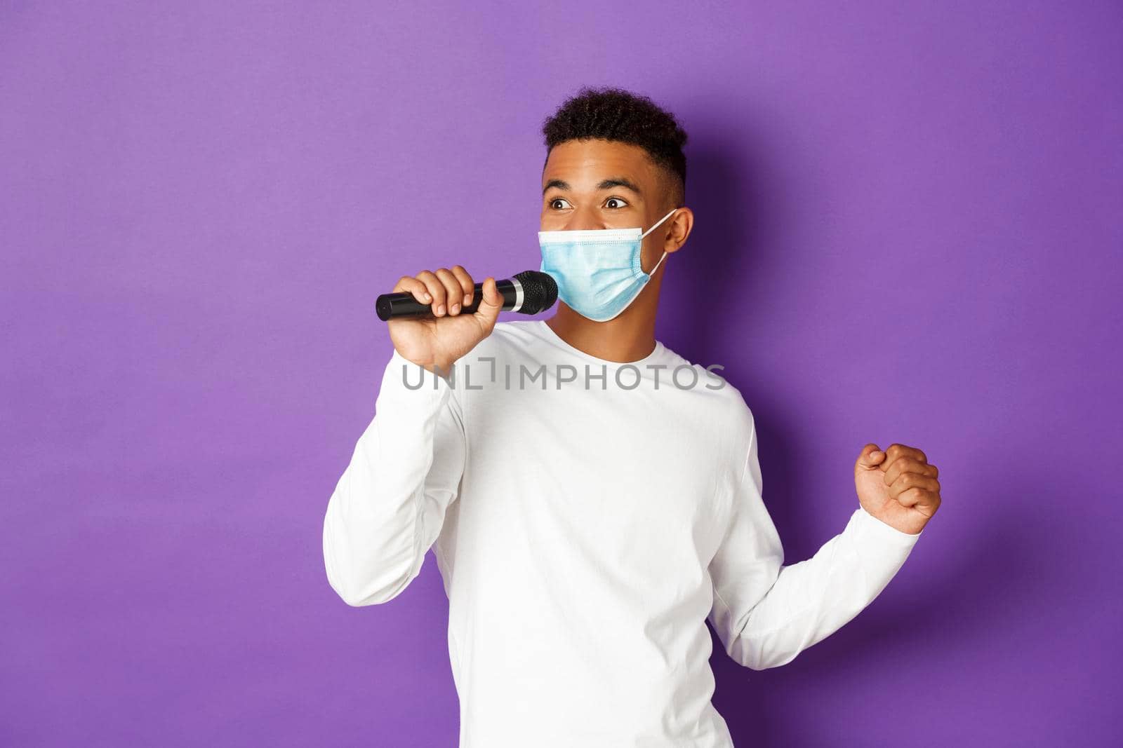 Concept of covid-19, pandemic and social distancing. Cheerful african-american guy having fun at karaoke, wearing medical mask and singing in microphone, standing over purple background.