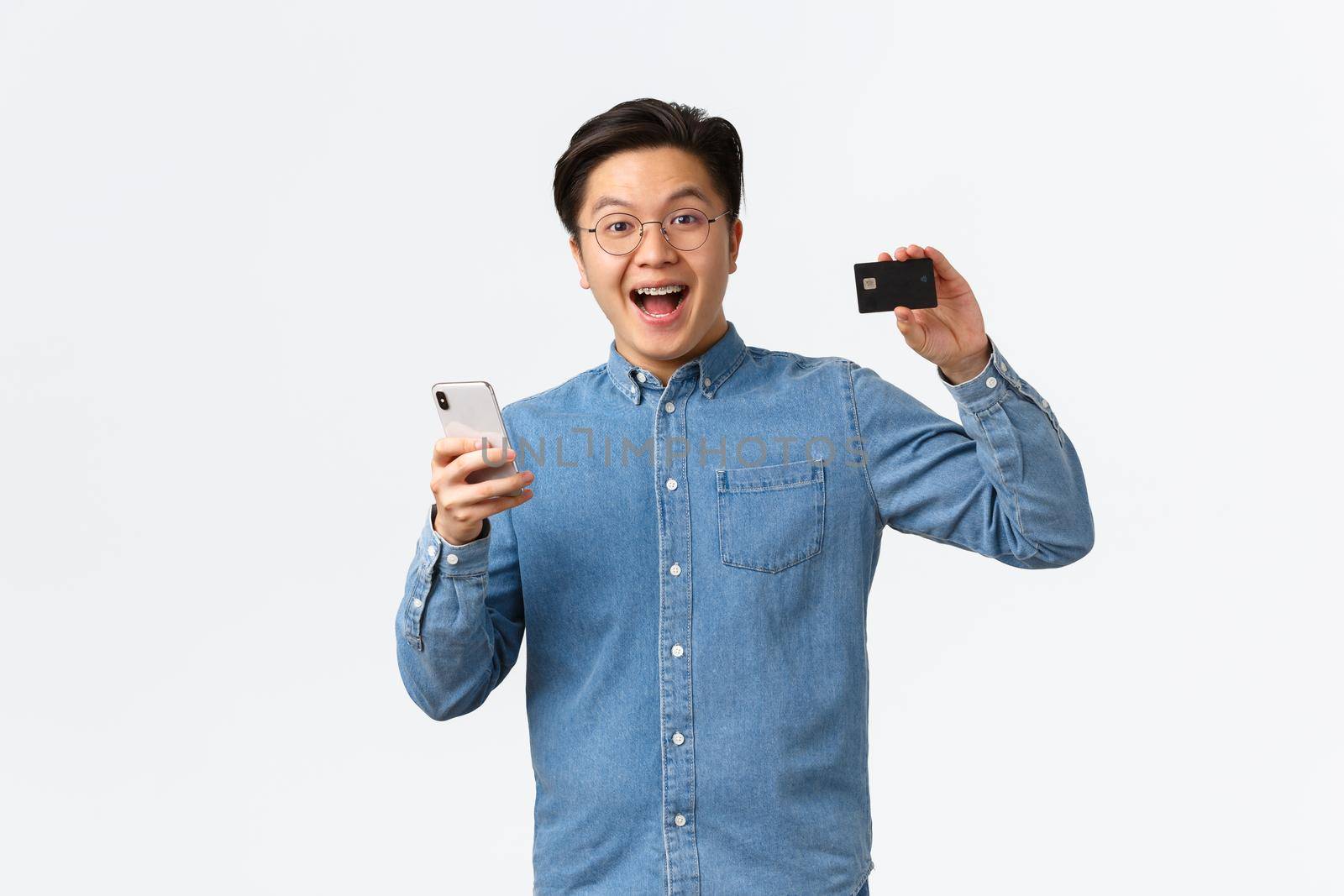 Excited and satisfied asian guy in glasses and casual clothes showing credit card with proud expression, paying for something in internet using smartphone, shopping online, white background by Benzoix