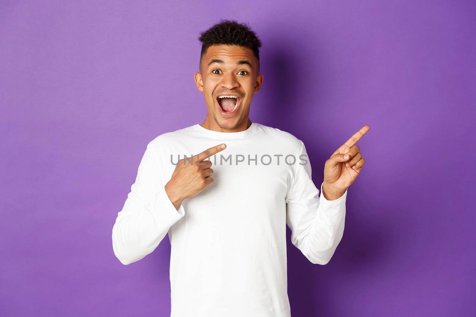 Portrait of handsome african-american guy in white sweatshirt, pointing fingers at upper right corner and smiling amazed, pointing fingers right at something cool, standing over purple background by Benzoix