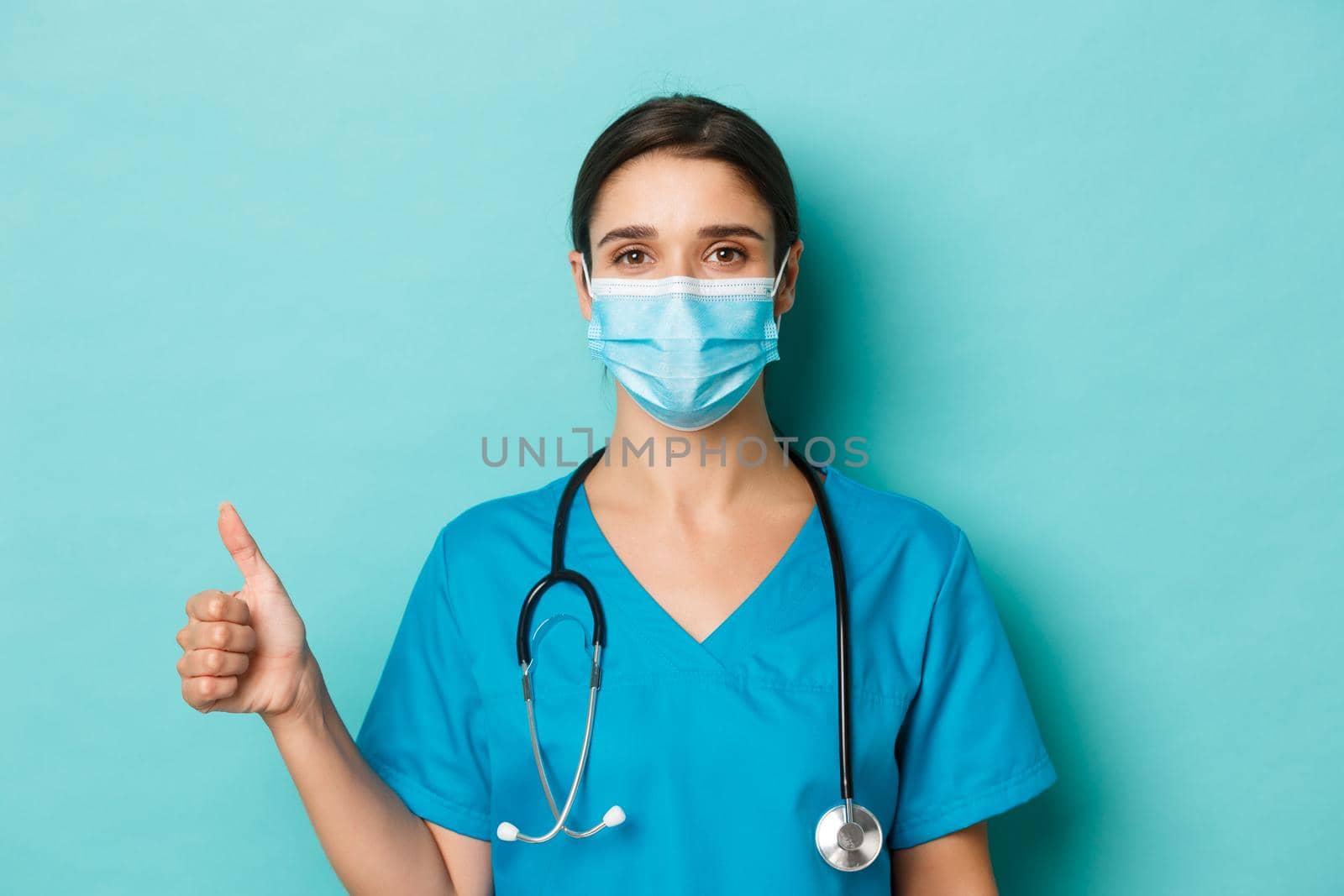 Concept of covid-19 and quarantine concept. Close-up of confident female doctor in medical mask and scrubs, holding stethoscope, showing thumbs-up, standing over blue background.