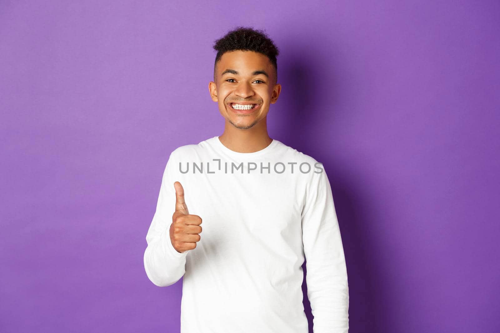 Image of handsome cheerful african-american young man, showing thumb-up and smiling pleased, praise good choice, looking satisfied, standing over purple background.