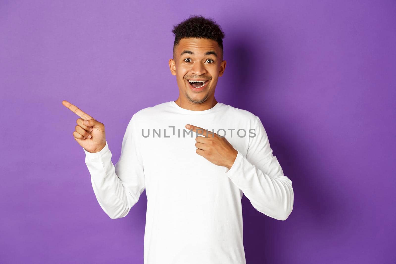 Image of excited and happy african-american guy pointing fingers at upper left corner, smiling amazed, showing cool advertisement, standing over purple background.