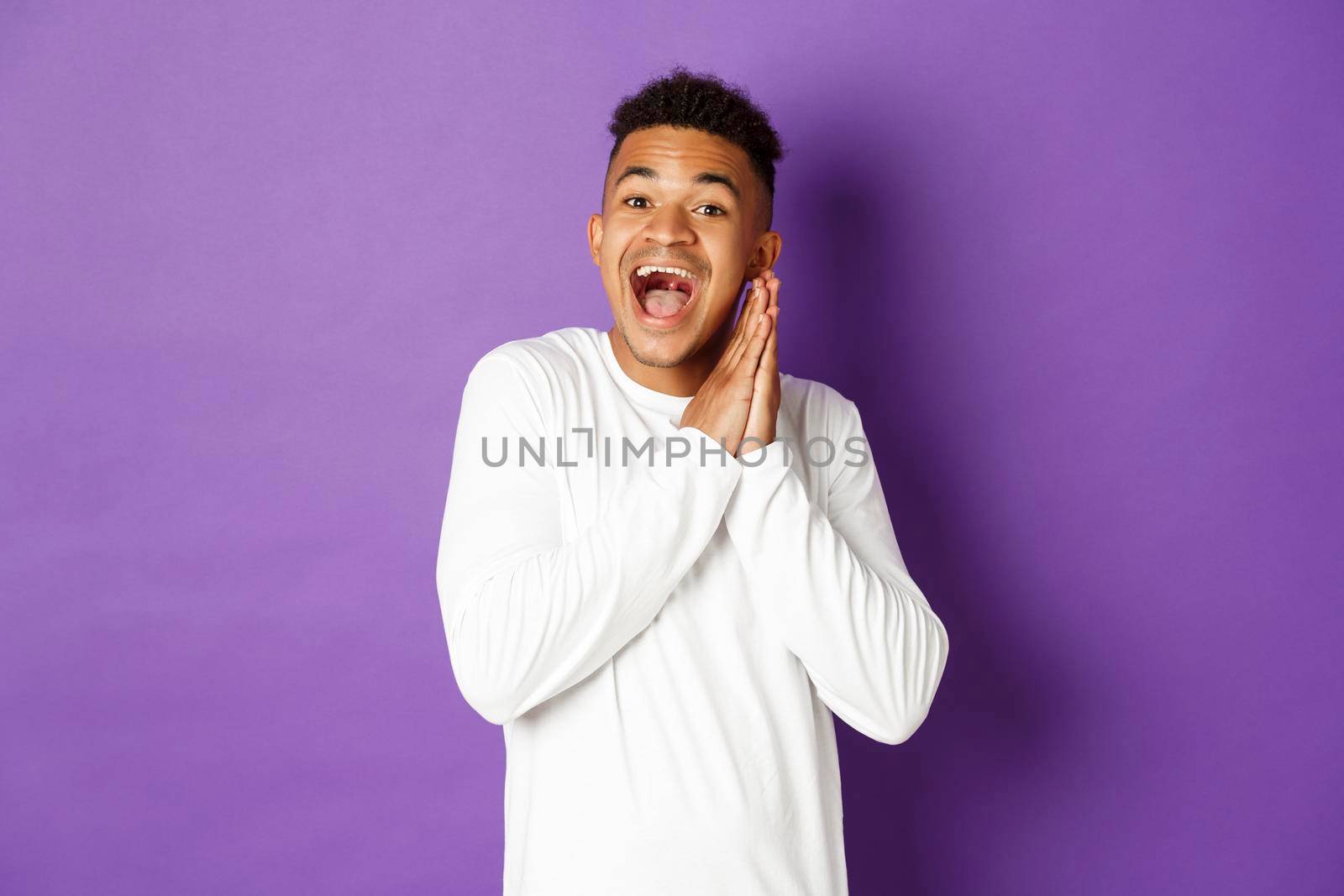 Image of excited and hopeful african-american queer guy, looking with anticipation and thrill at something, clap hands and smiling happy, standing over purple background by Benzoix