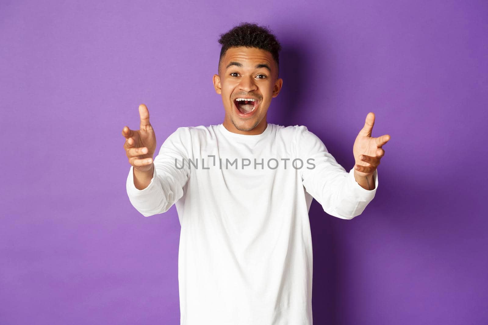 Portrait of handsome african-american young man, reaching hands forward to receive something, looking amazed and happy, standing over purple background.