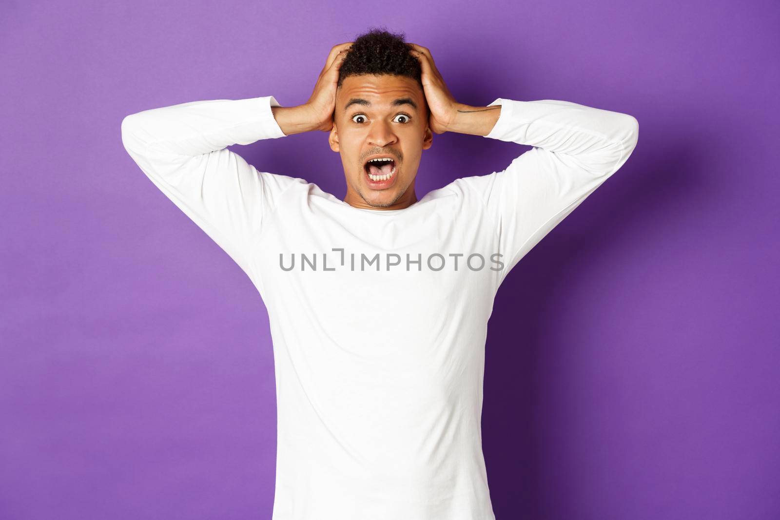 Image of handsome african-american man in panic, holding hands on head and looking nervous at camera, standing alarmed and scared over purple background.