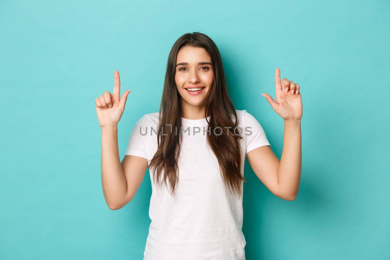 Portrait of happy beautiful woman in white t-shirt, smiling and pointing fingers up, showing logo, standing over blue background by Benzoix