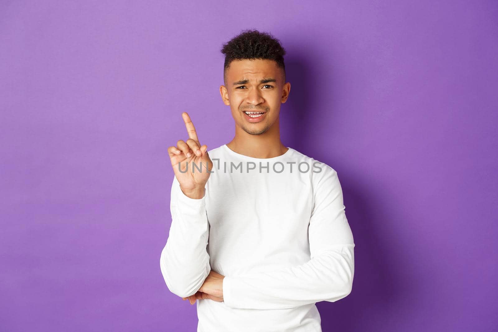 Image of skeptical young african-american man in white sweatshirt, forbid something, frowning disappointed and shaking finger in disapproval, dislike idea, standing over purple background by Benzoix