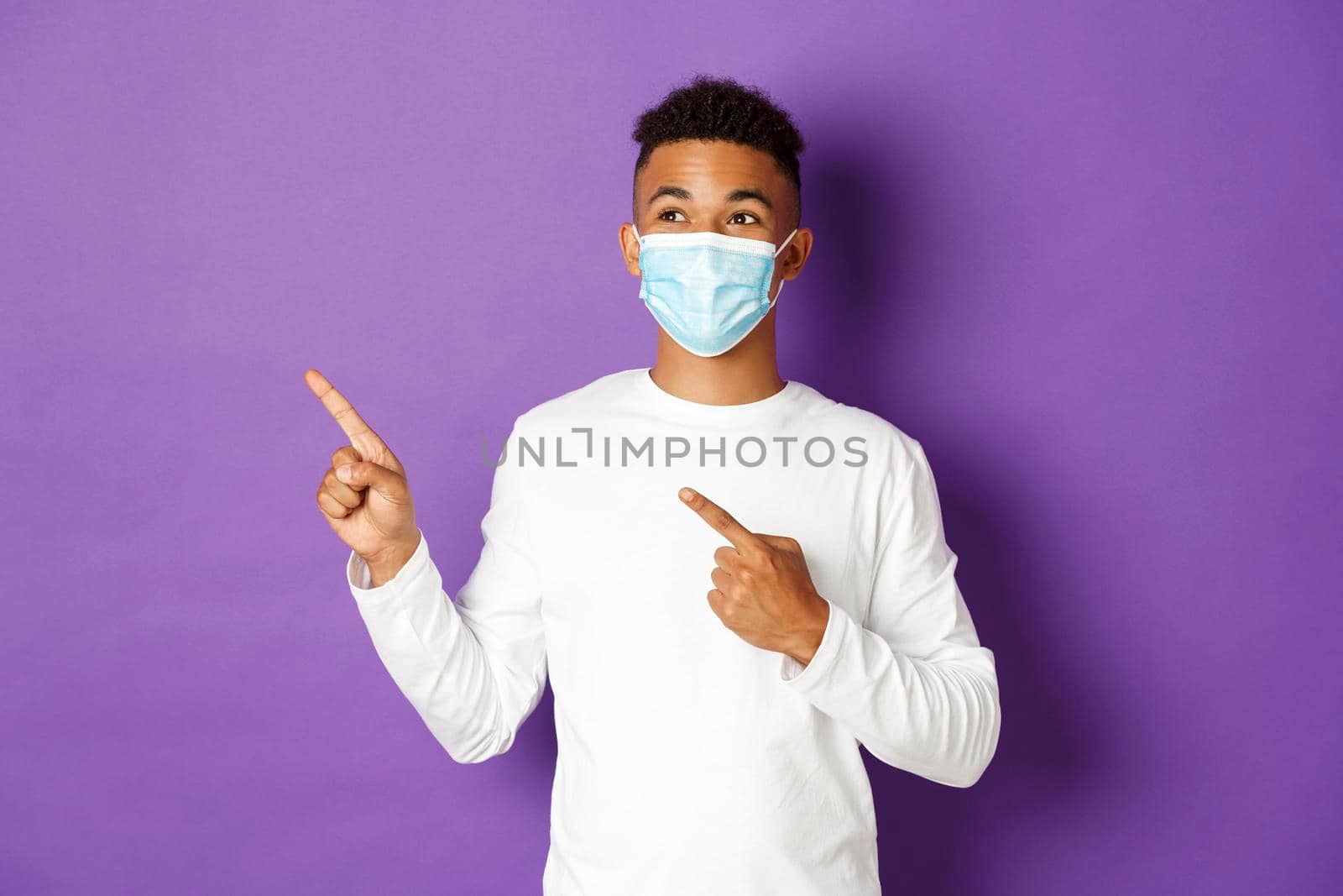 Concept of coronavirus, quarantine and lifestyle. Handsome young african-american man in medical mask, pointing and looking at upper left corner, showing advertisement, posing over purple background.