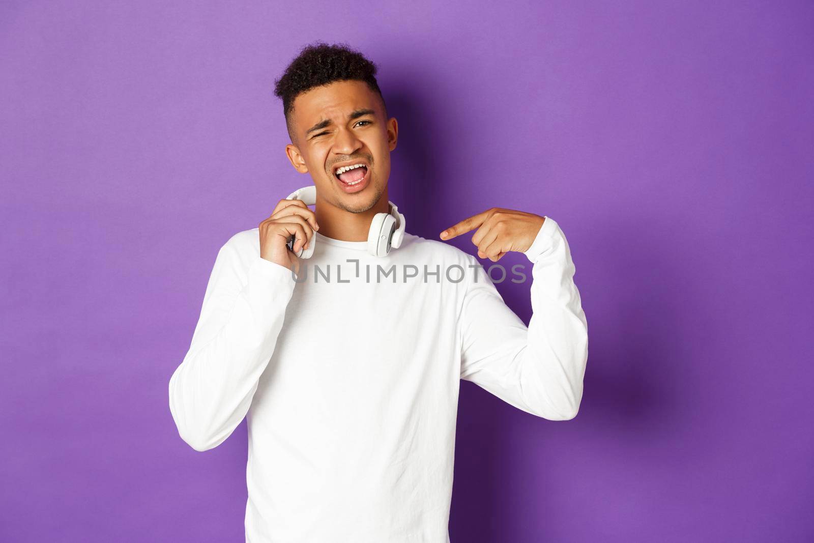 Image of sassy african-american guy, pointing at headphones and praising awesome song, standing over purple background.