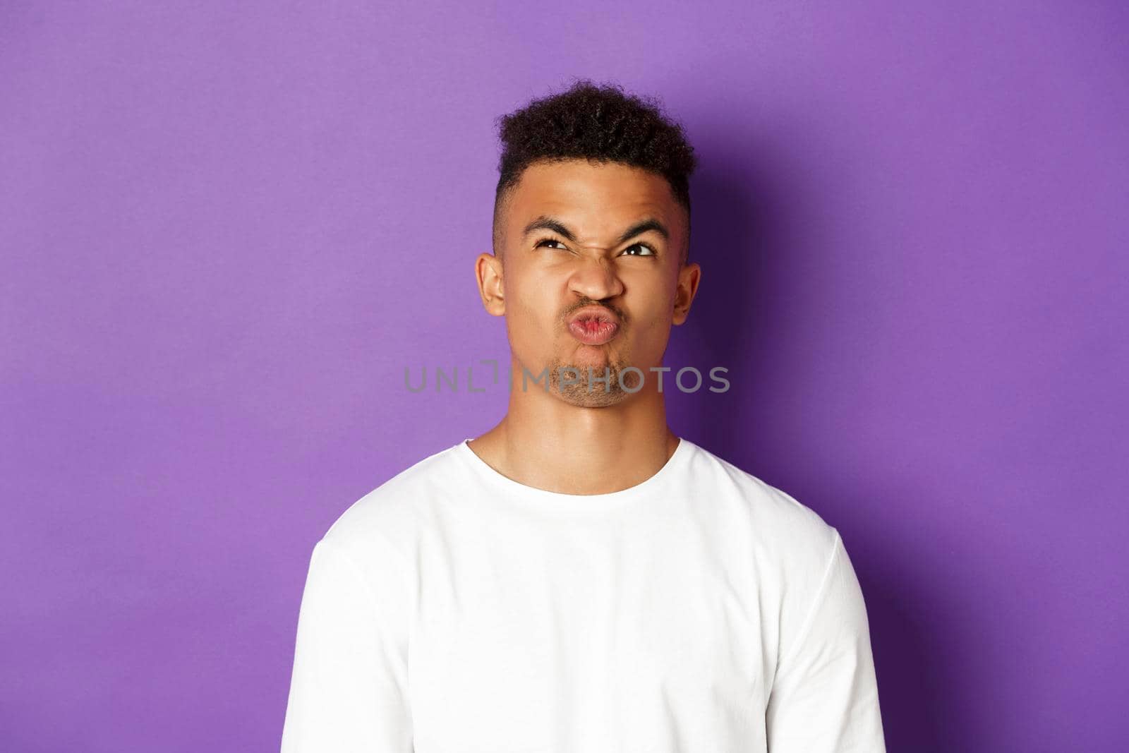 Close-up of troubled young african-american guy, looking up and grimacing perplexed, having hard choice, standing over purple background.