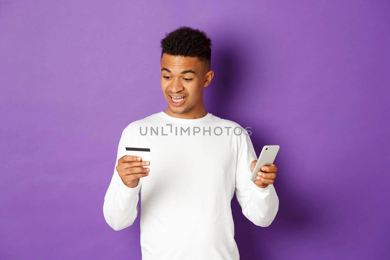 Portrait of african-american guy looking amazed at credit card, shopping online on smartphone app, standing over purple background.