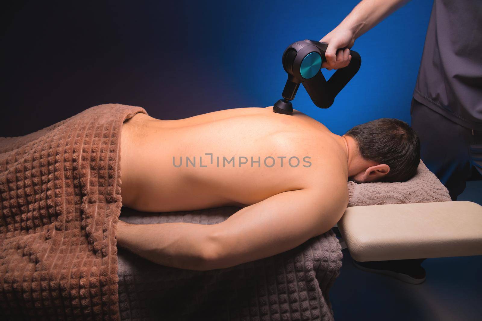 Physiotherapy of the upper back with a percussion massager. Therapist kneads the patient's upper back.