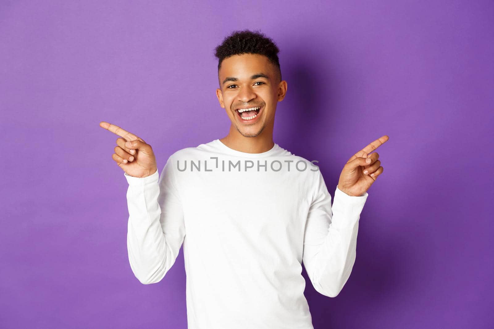 Image of cheerful african-american guy in white sweatshirt, showing two variants, pointing fingers sideways at copy space and smiling, demonstrate products, purple background.