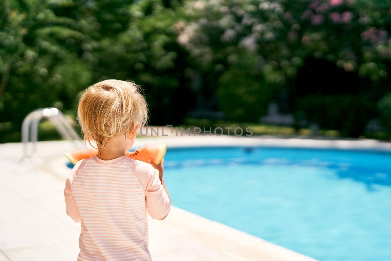 Little baby stands by the pool, holding a watermelon in his hands. Back view. High quality photo