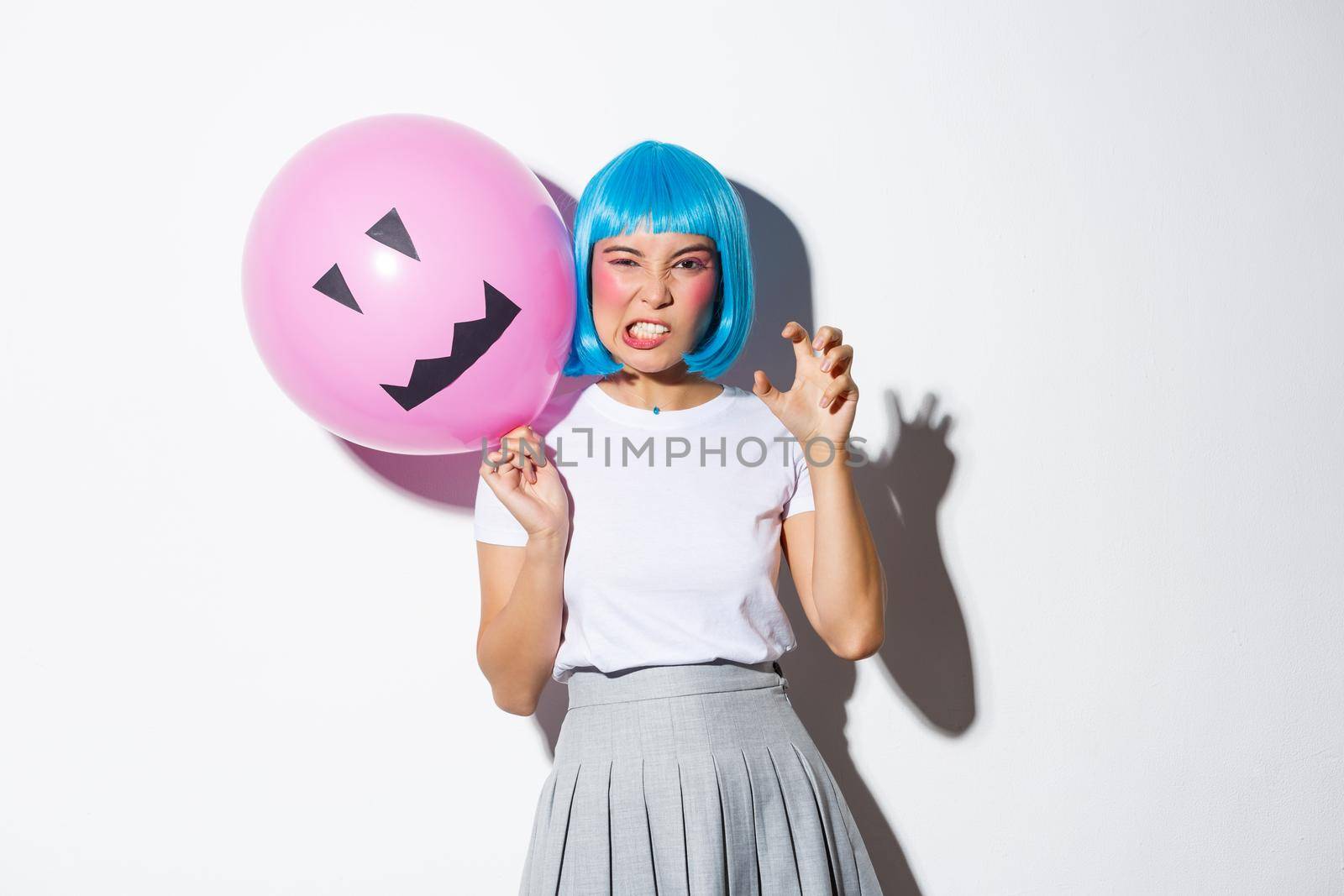 Portrait of funny asian girl trying to scare someone on halloween, wearing blue wig and holding pink balloon with scary face.