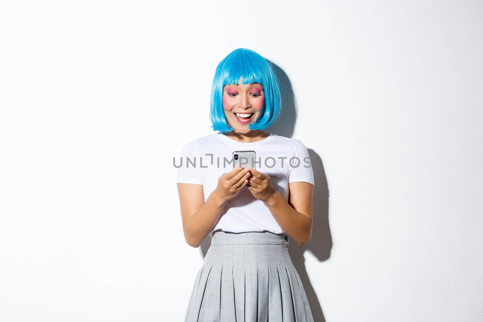 Image of attractive asian girl looking at mobile phone screen with happy and surprised face, standing in blue anime wig and halloween costume.