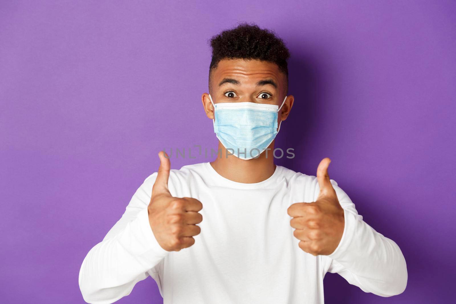 Concept of coronavirus, quarantine and social distancing. Close-up of young african-american guy in medical mask, looking excited and recommending something, showing thumbs-up in approval.
