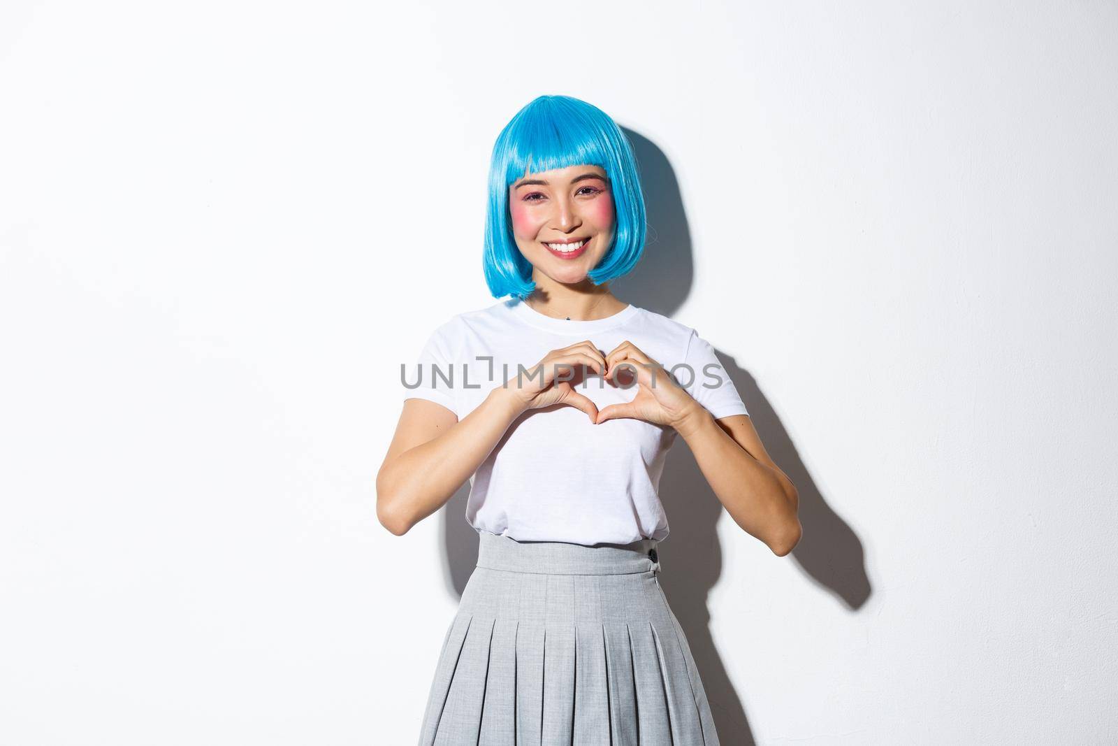 Lovely asian woman in blue party wig and colorful makeup showing heart gesture, smiling with gratitude, standing over white background.