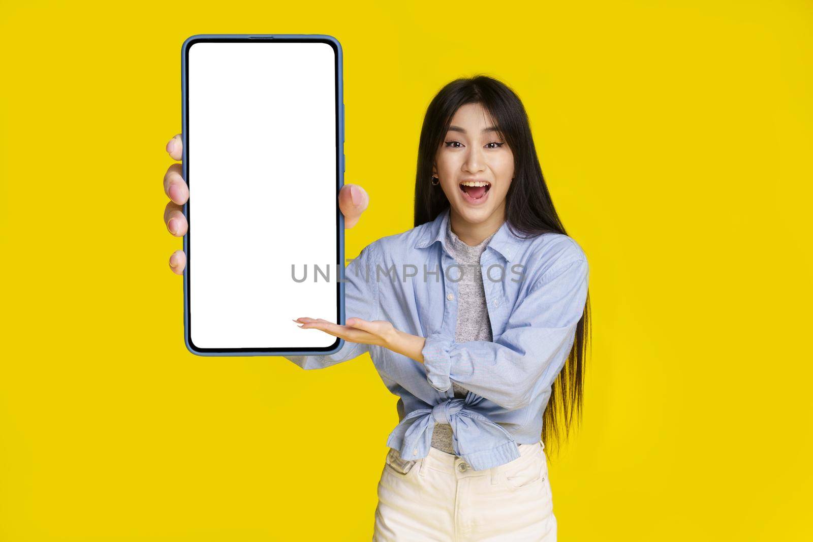 Big smartphone in hand of beautiful asian girl with white screen happy to introduce new app, game, win, isolated on yellow background. Mobile app advertisement product placement by LipikStockMedia