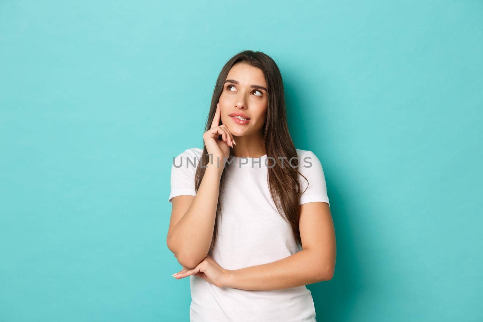 Portrait of thoughtful pretty girl in white t-shirt, looking at upper left corner and thinking, making decision, standing over blue background.