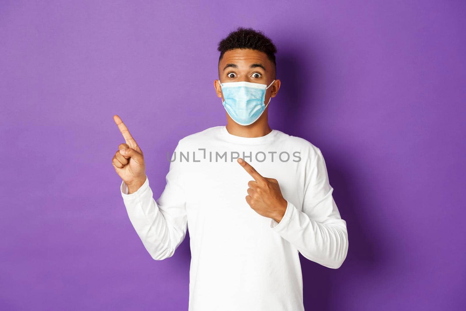 Concept of coronavirus, quarantine and lifestyle. Amazed african-american man in medical mask showing advertisement, pointing at upper left corner and smiling, standing over purple background.