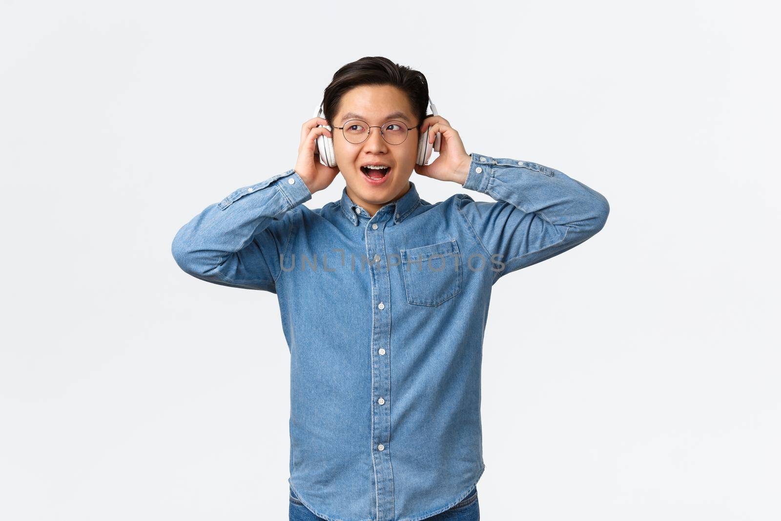 Lifestyle, leisure and technology concept. Smiling amazed asian guy in glasses, trying new wireless headphones, listening music with impressed and pleased face, standing white background by Benzoix