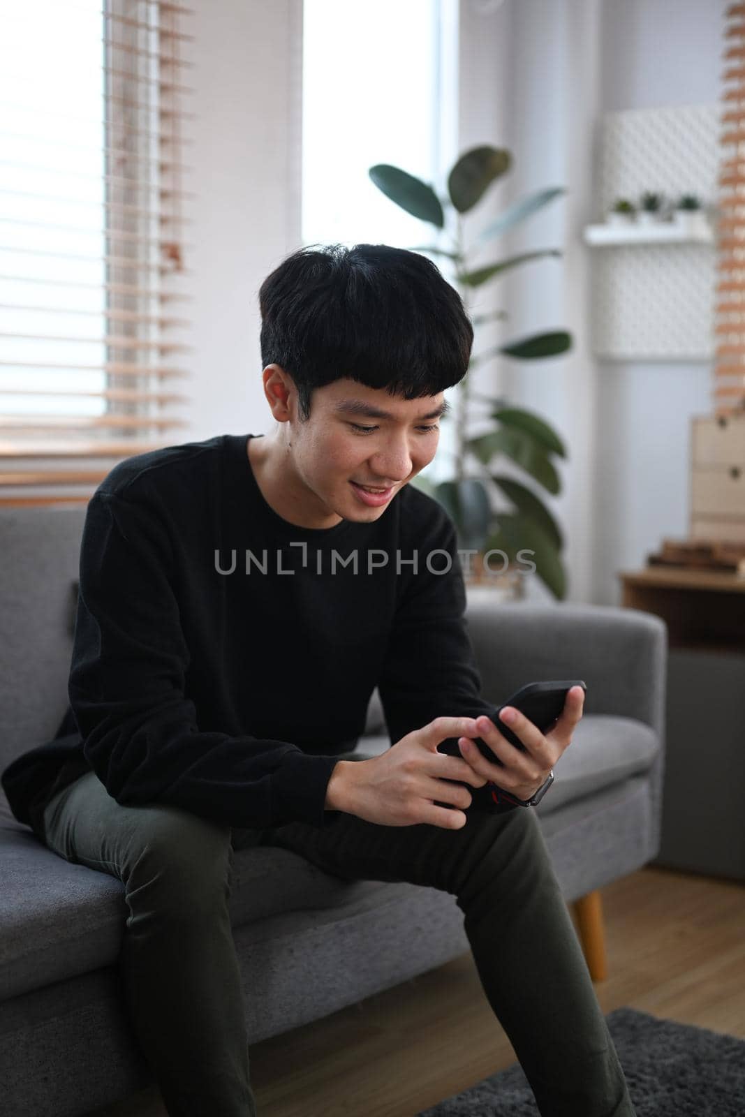 Smiling young asian man resting on couch in cozy living room and using mobile phone by prathanchorruangsak