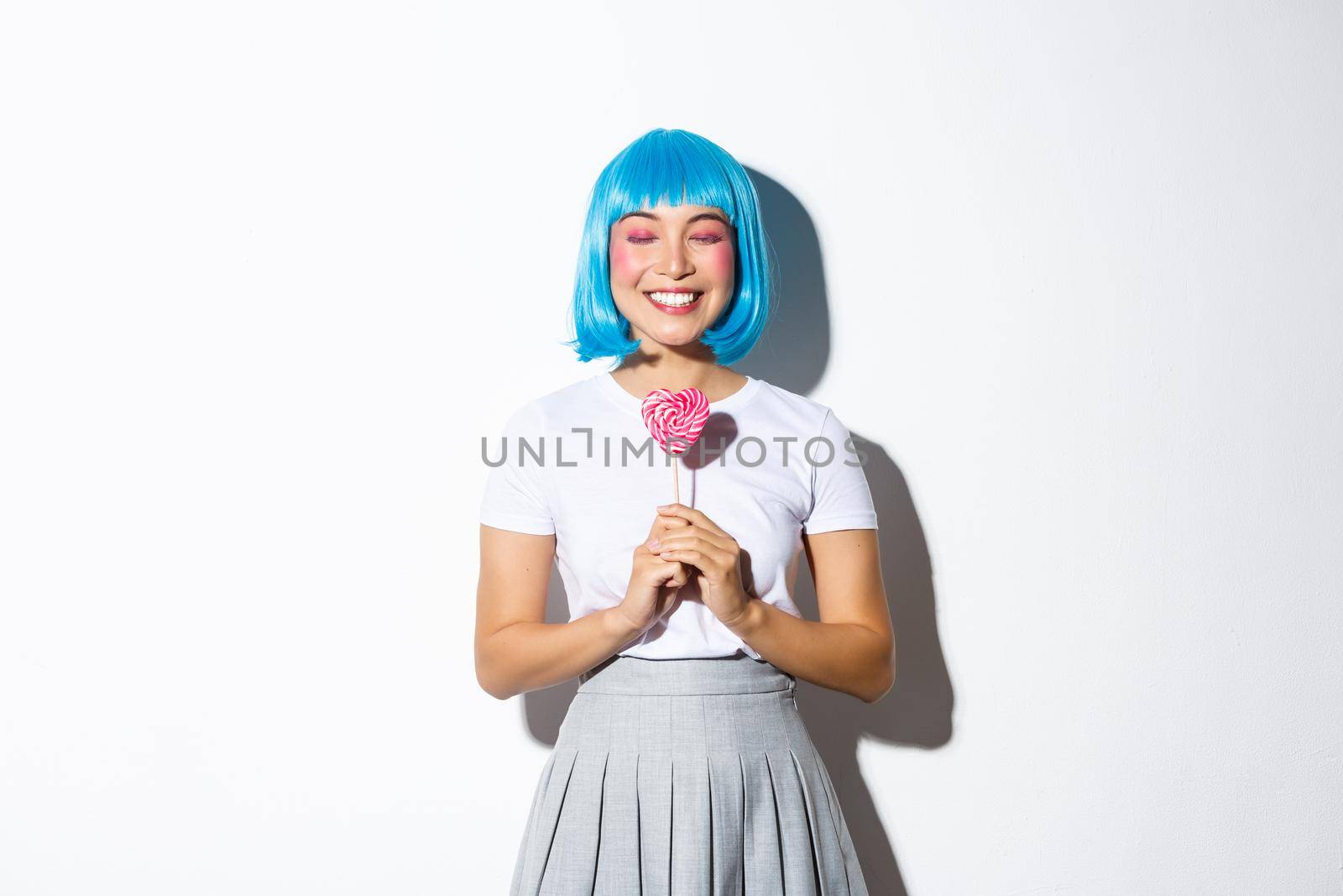Image of happy asian girl in blue short wig standing with eyes closed and dreamy smile, holding heart-shaped candy, standing over white background.