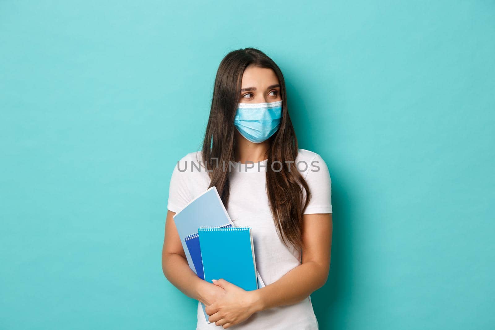 Concept of coronavirus, health and social distancing. Portrait of female student attend classes in medical mask, looking right at copy space, holding notebooks, standing over blue background by Benzoix