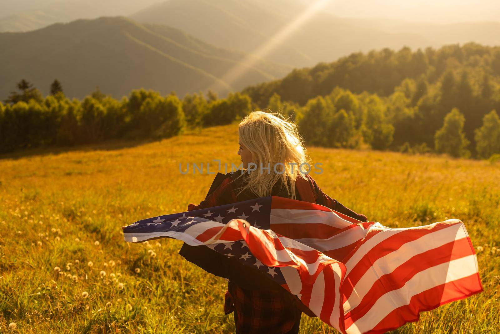Young happy american woman with long hair holding waving on wind USA national flag on her sholders relaxing outdoors enjoying warm summer day.