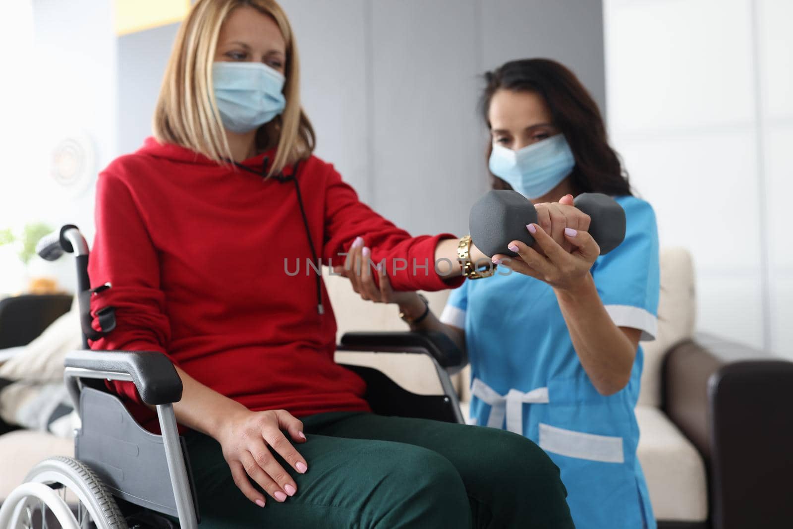 Doctor or nurse is training with woman with dumbbell both in protective masks at home. Rehabilitation after injury or stroke concept