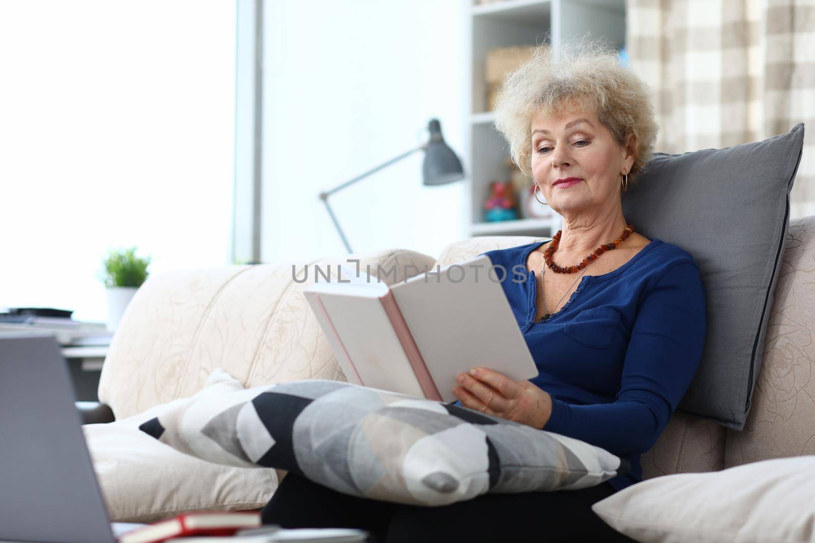 Elderly woman is reading book at home on couch. Home leisure pensioners concept