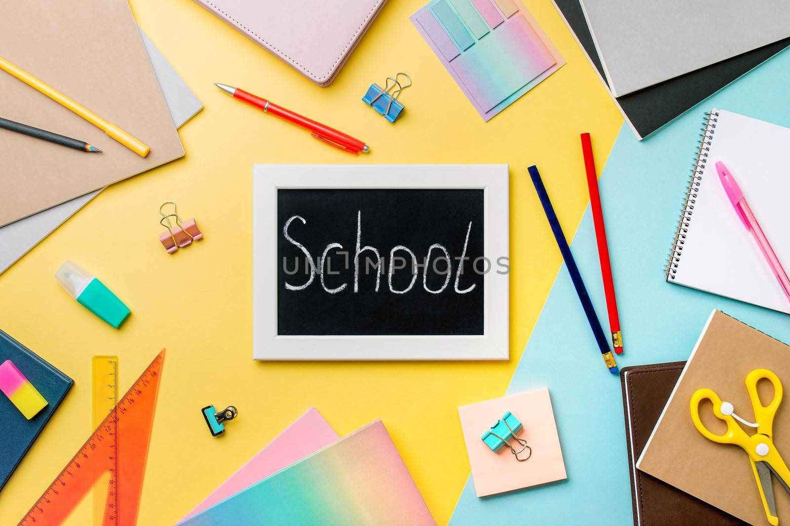 Blackboard with letters school made with chalk. School supplies, pencils, scissors and colored books on yellow and blue background. Top view or flat lay. Back to school concept