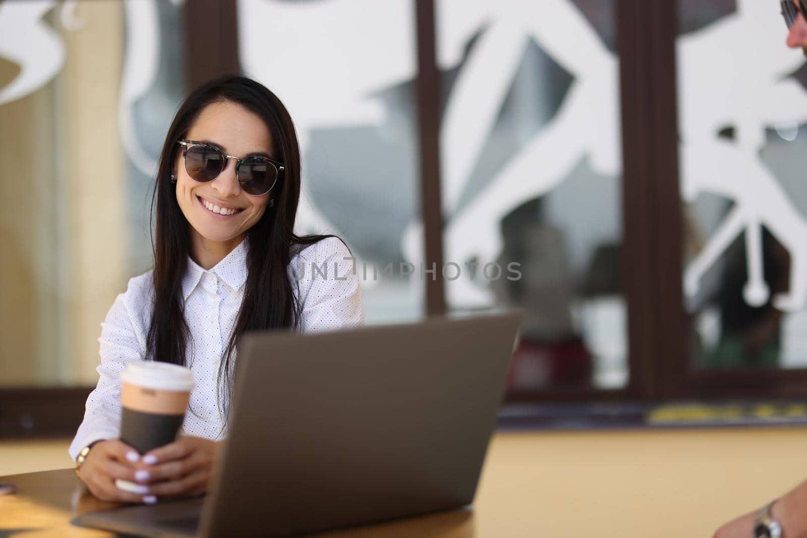 Smiling sun woman holding coffee and working on laptop in cafe. Blogger and remote work concept