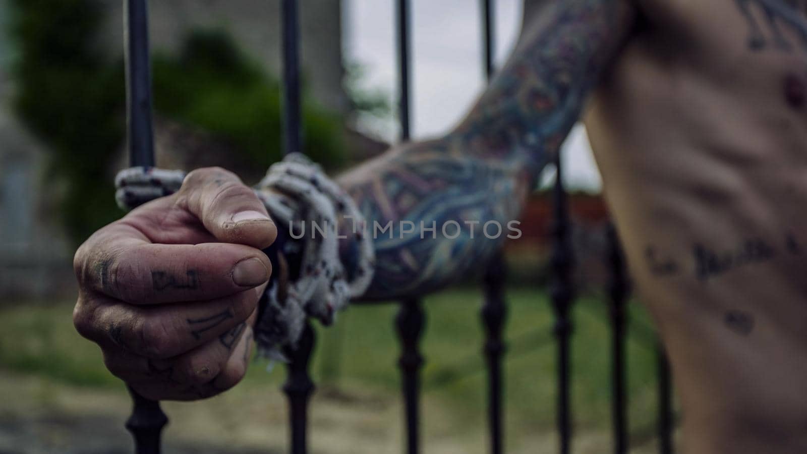 Details of tattooed hands of a man tied to grills behind him struggles to escape