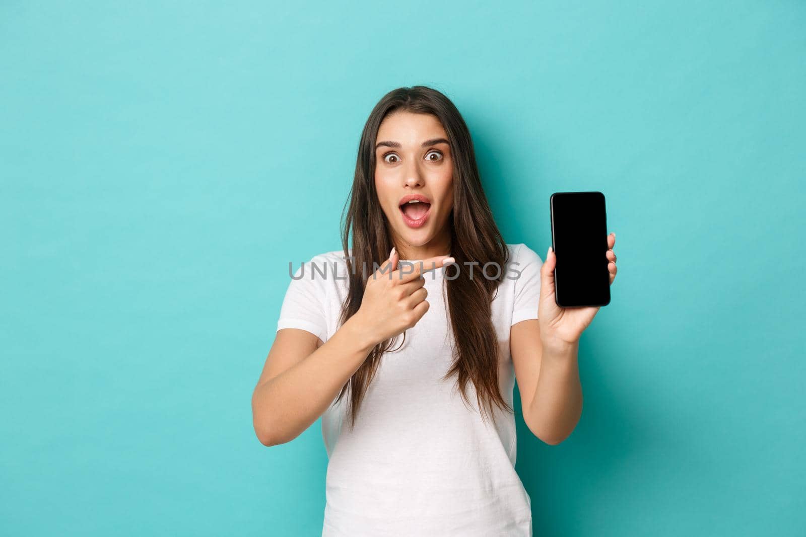 Portrait of amazed pretty woman in white t-shirt, pointing finger at mobile phone screen, standing excited over blue background.