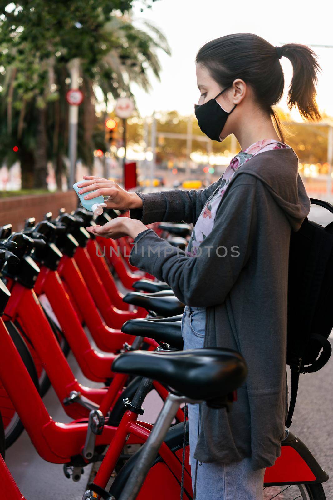 vertical photo of a young girl with protective mask cleaning her hands with disinfectant gel next to an urban bicycle rental parking, concept of protection against covid and sustainable mobility