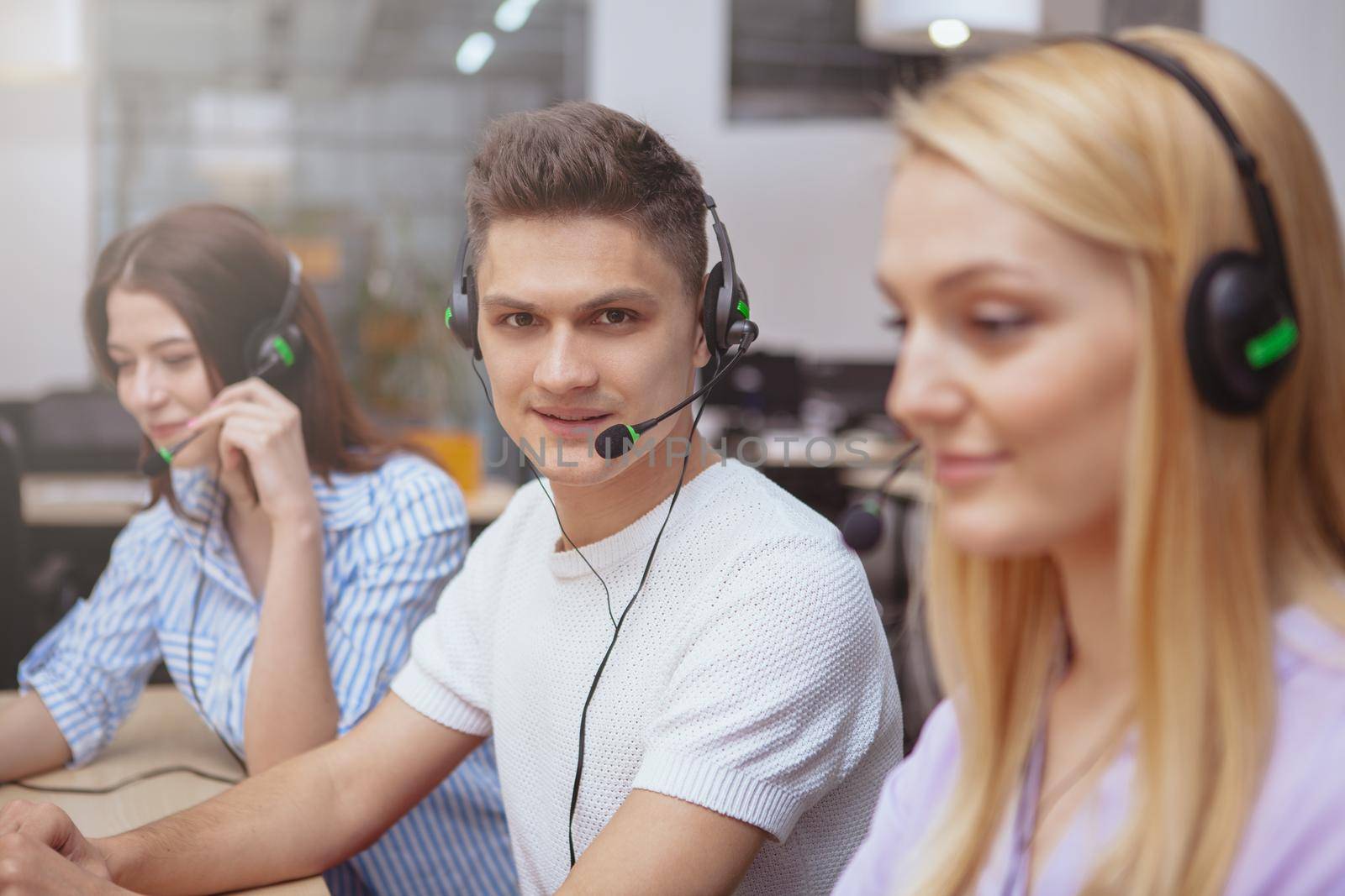 Young people working at call center, wearing headsets with microphones, answering calls. Handsome cheerful male customer support operator smiling to the camera, while working on laptop