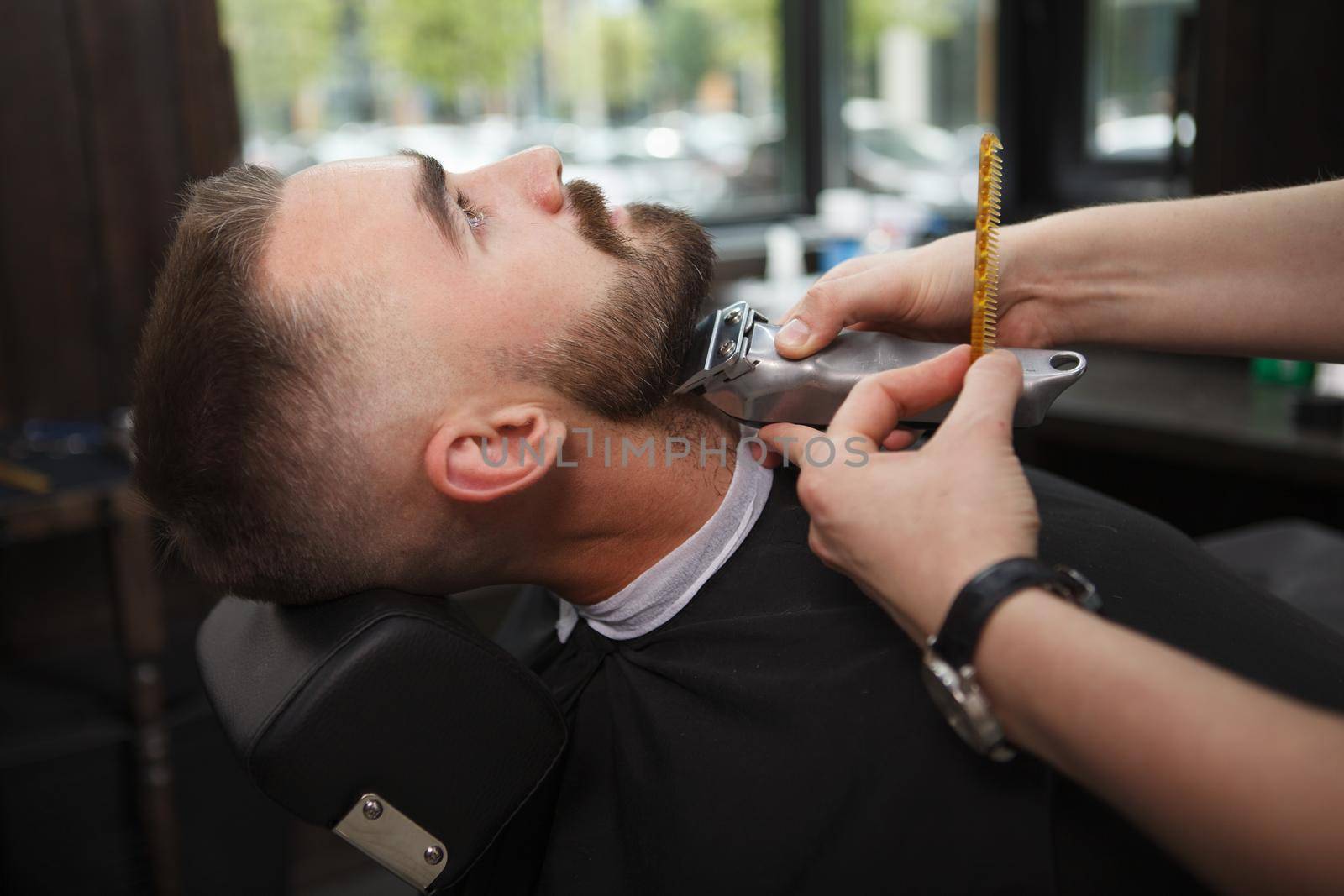 Male client relaxing while professional barber trimming his beard