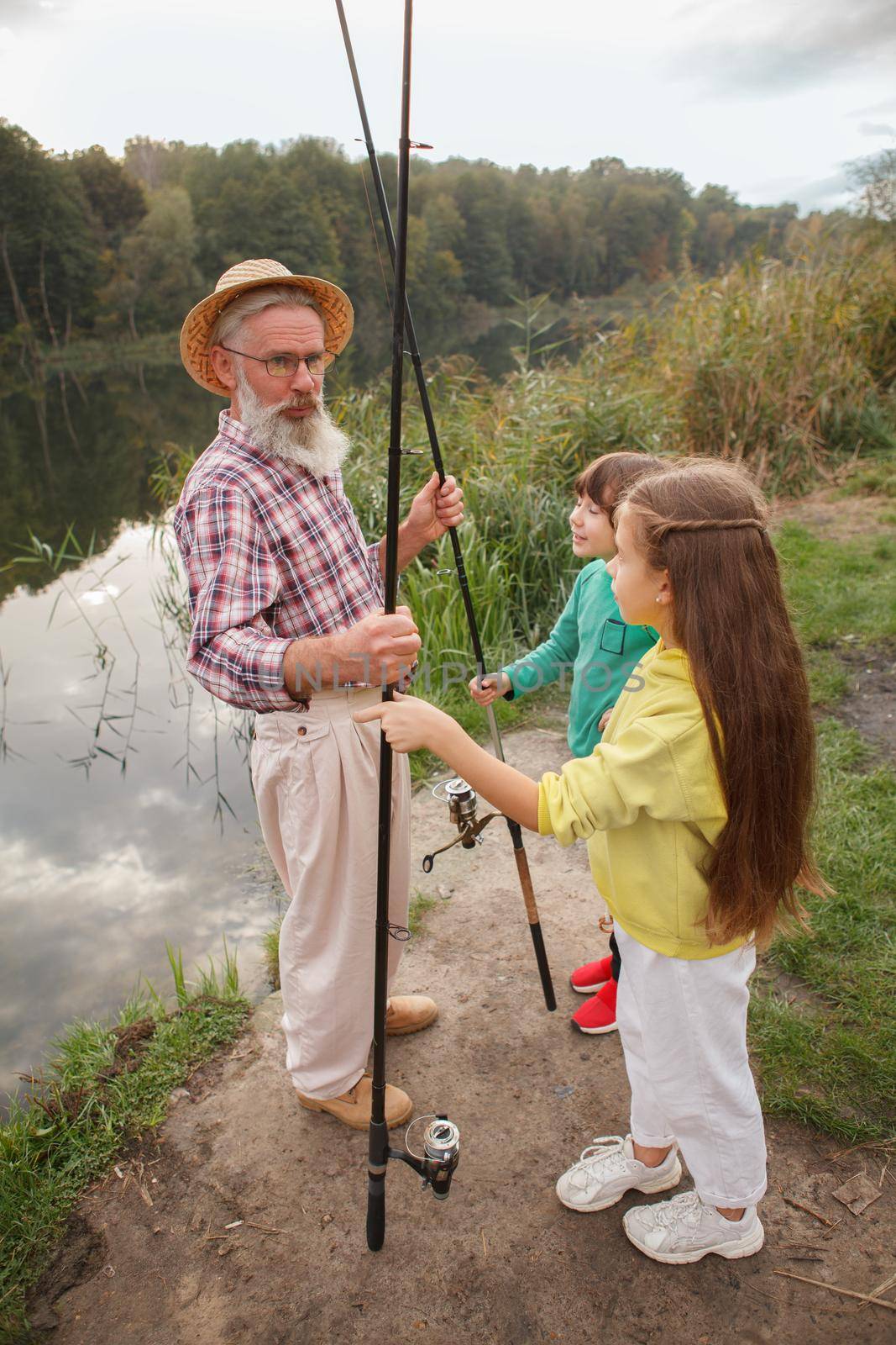 Vertical full length shot of senior man and his two grandchild preparing to fish on the local lake at countryside