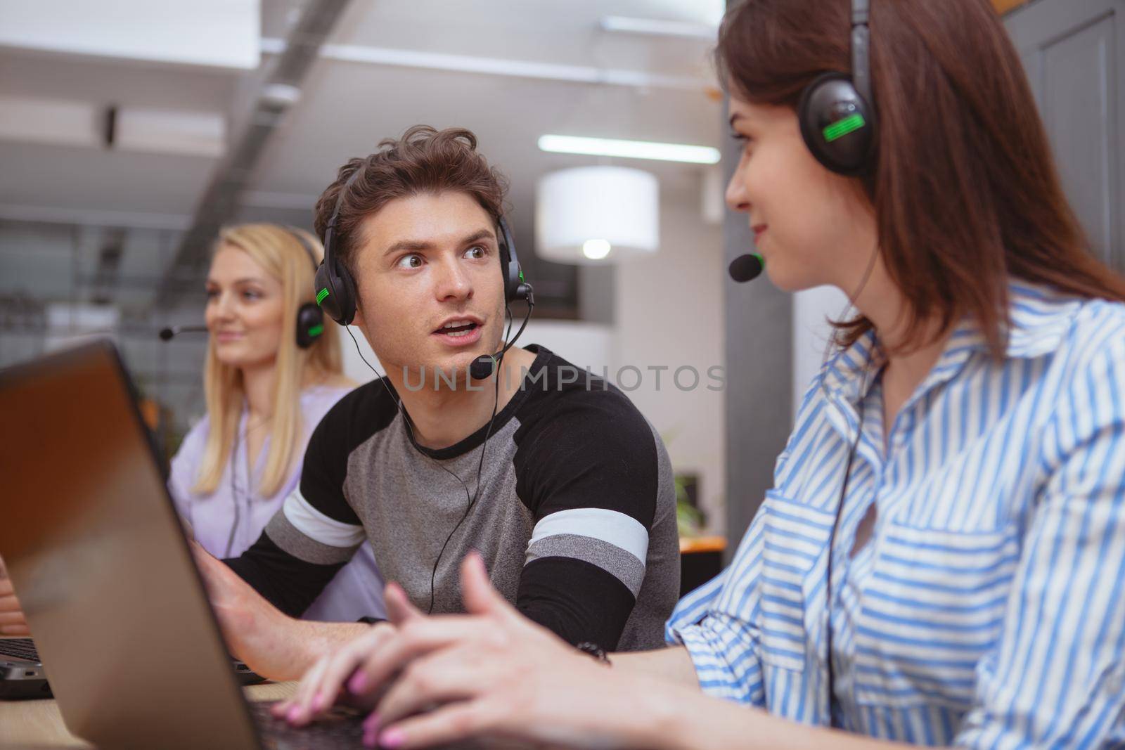 Young people working at customer support service, wearing headsets, working on computers. Attractive male call center operator talking to a colleague