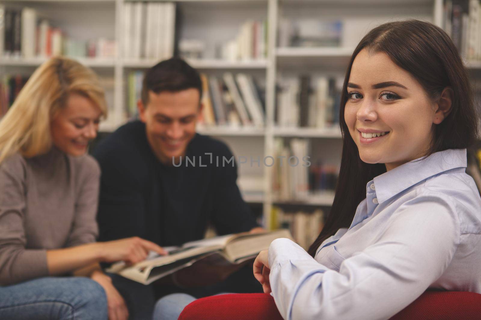 Lovely happy female student smiling to the camera, sitting relaxed at the college library, her classmates reading on the background. Cheerful woman resting in an armchair at the campus library, copy space