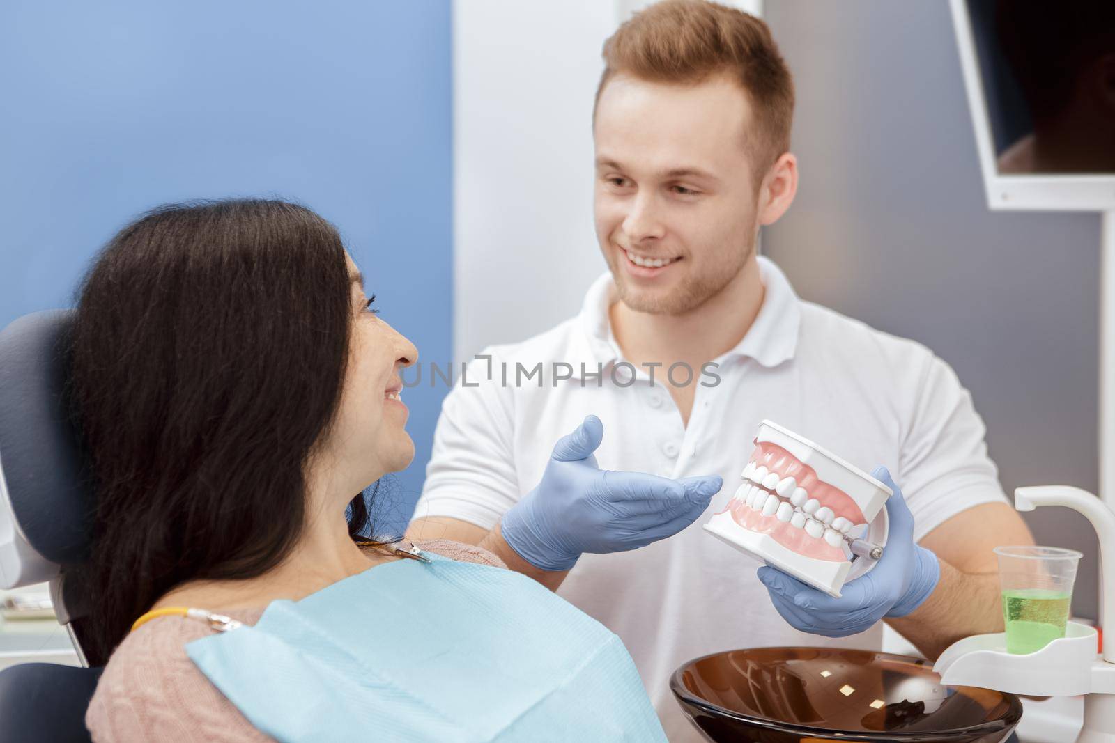 Teeth care. Handsome young professional dentist explaining his senior patient how to care after teeth showing a teeth mold working at dental clinic professional communication medical service concept