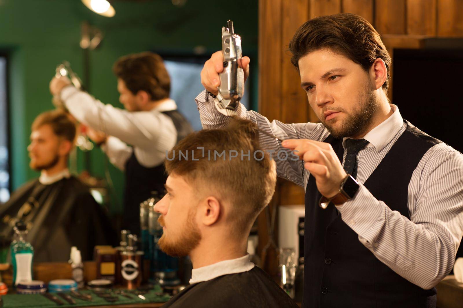 Attractive bearded professional barber giving his male client a haircut using electric trimmer clipper professionalism styling hairdressing occupation career job traditional barbering barbershop service.