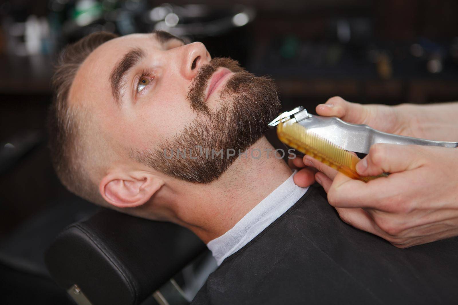 Close up of a handsome bearded man at the barbershop having his beard trimmed with electric clipper