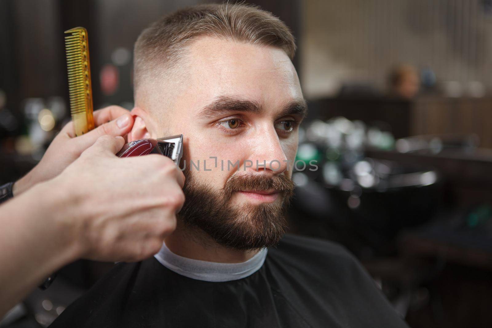 Close up of a handsome bearded man getting his hair trimmed by professional barber