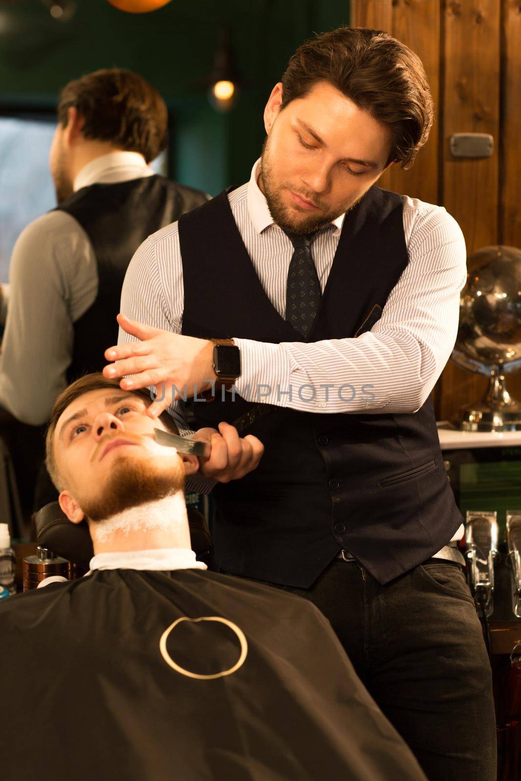 Vertical shot of a handsome professional barber working shaving his client at the barbershop occupation razor equipment barbering job lifestyle hipster traditional hairdresser stylist.