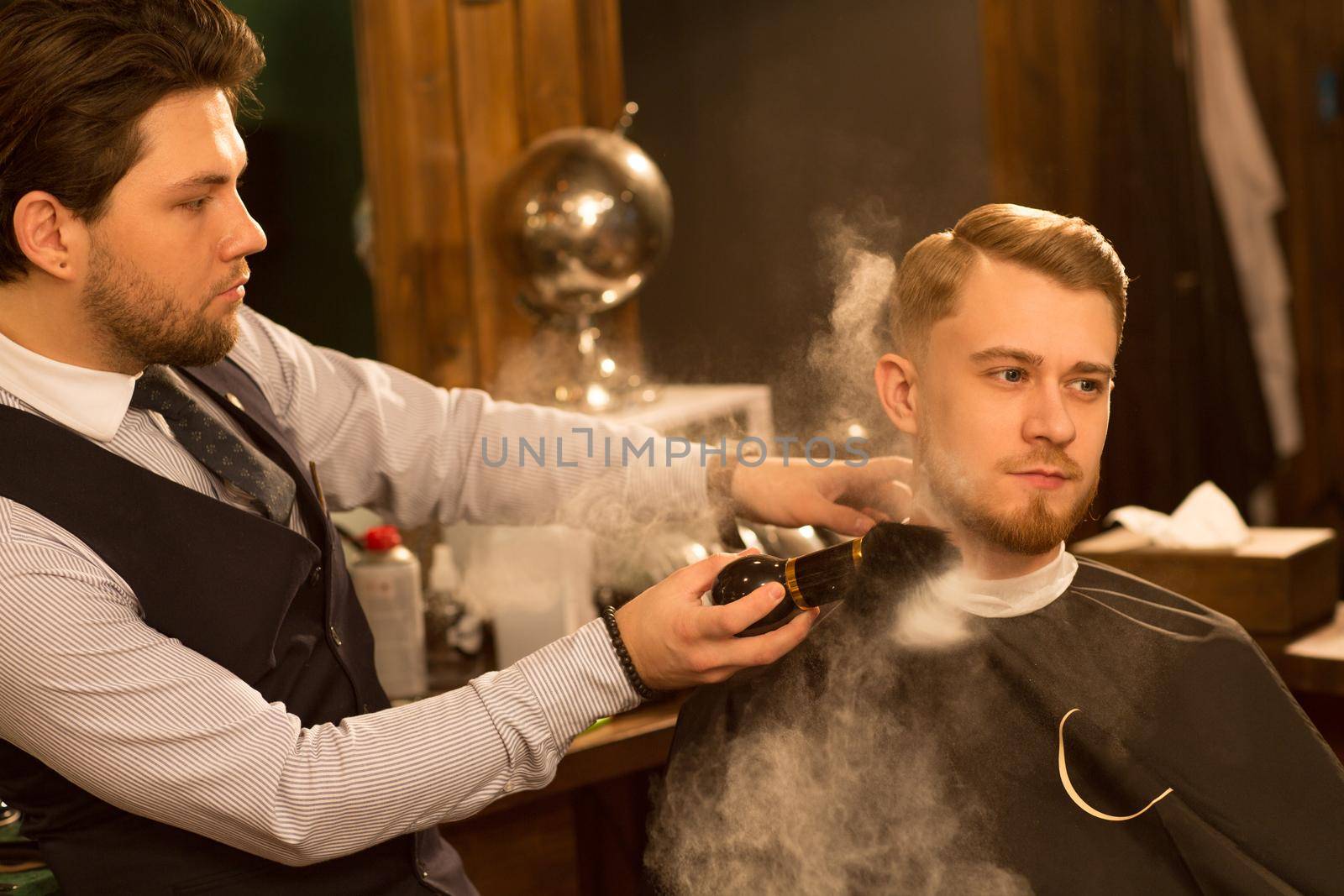 Handsome young man looking away sitting in the barber chair professional barber using talcum powder after shaving his client work employee service consumerism occupation job lifestyle hipster skincare
